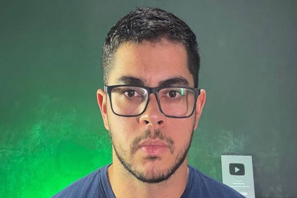 Homem jovem de cabelo curto e barba aparada usa óculos de armação preta e camiseta azul escura. Ele está posicionado centralmente contra um fundo cinza com iluminação verde à esquerda e um troféu do YouTube desfocado à direita.