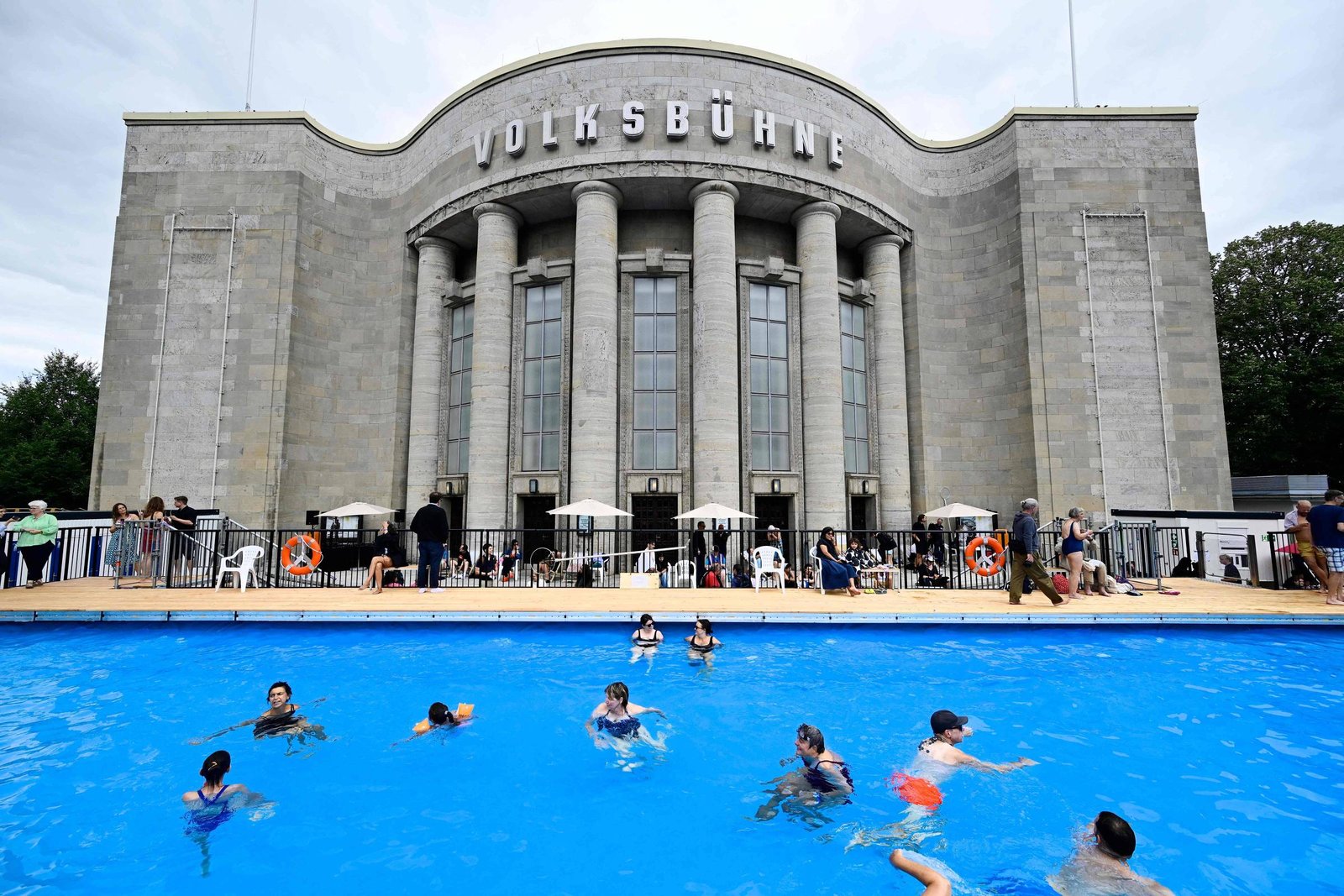 Pessoas nadam e brincam em piscina azul em primeiro plano, com o teatro Volksbühne de pedra cinza ao fundo. O edifício tem colunas altas e o nome
