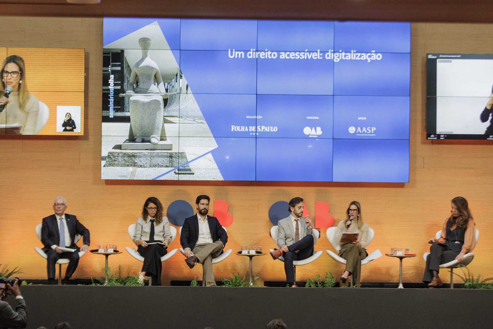 Seis pessoas sentadas em cadeiras em um palco com fundo laranja, participando de um painel de debate. Ao fundo, tela grande exibe imagem de estátua e texto em francês. Público assiste em cadeiras dispostas em fileiras.