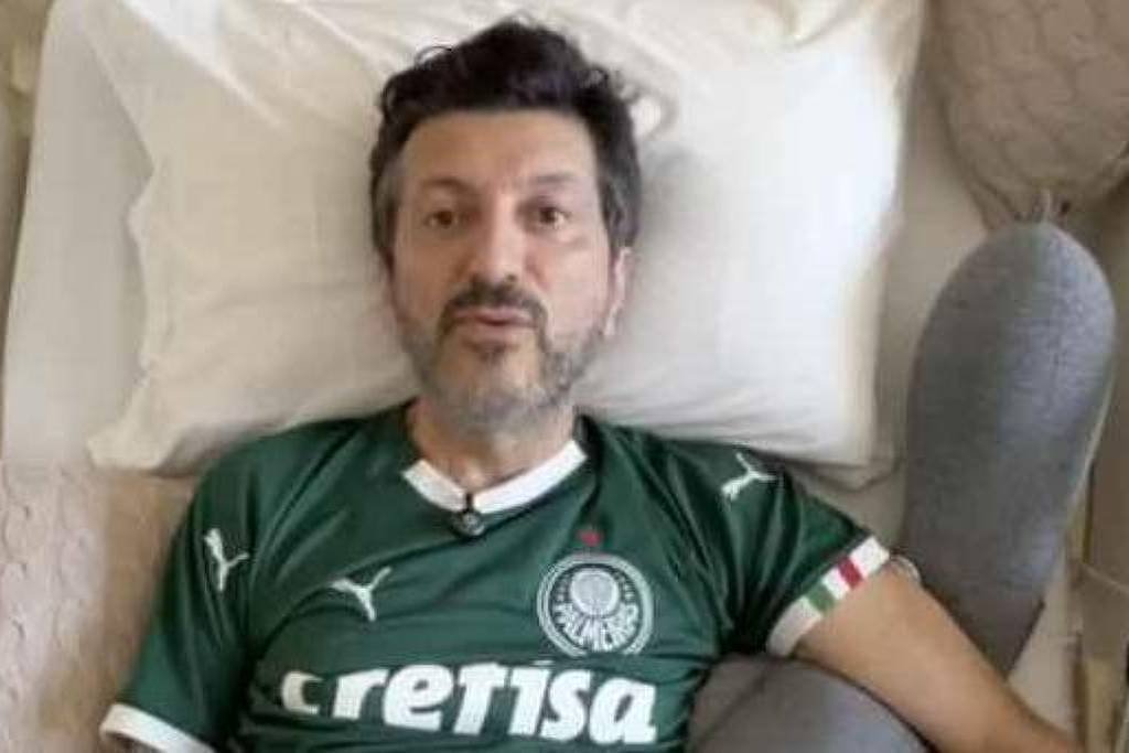 Homem de meia-idade deitado em cama branca, usando camisa verde do Palmeiras com logo da Crefisa. Travesseiro branco e almofada bege ao lado.