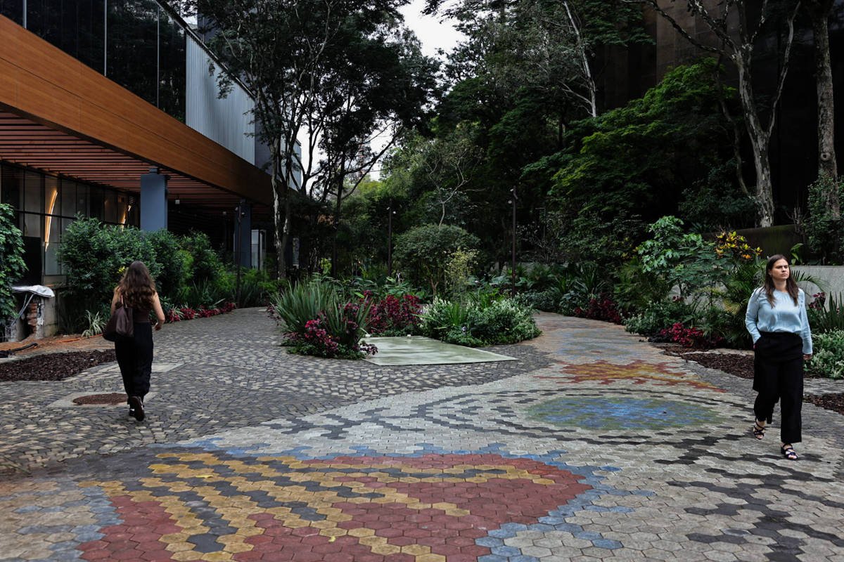 Duas mulheres caminham em caminhos de mosaico colorido em área externa com vegetação densa e edifícios ao fundo. O caminho se divide em duas direções, cercado por plantas e árvores.