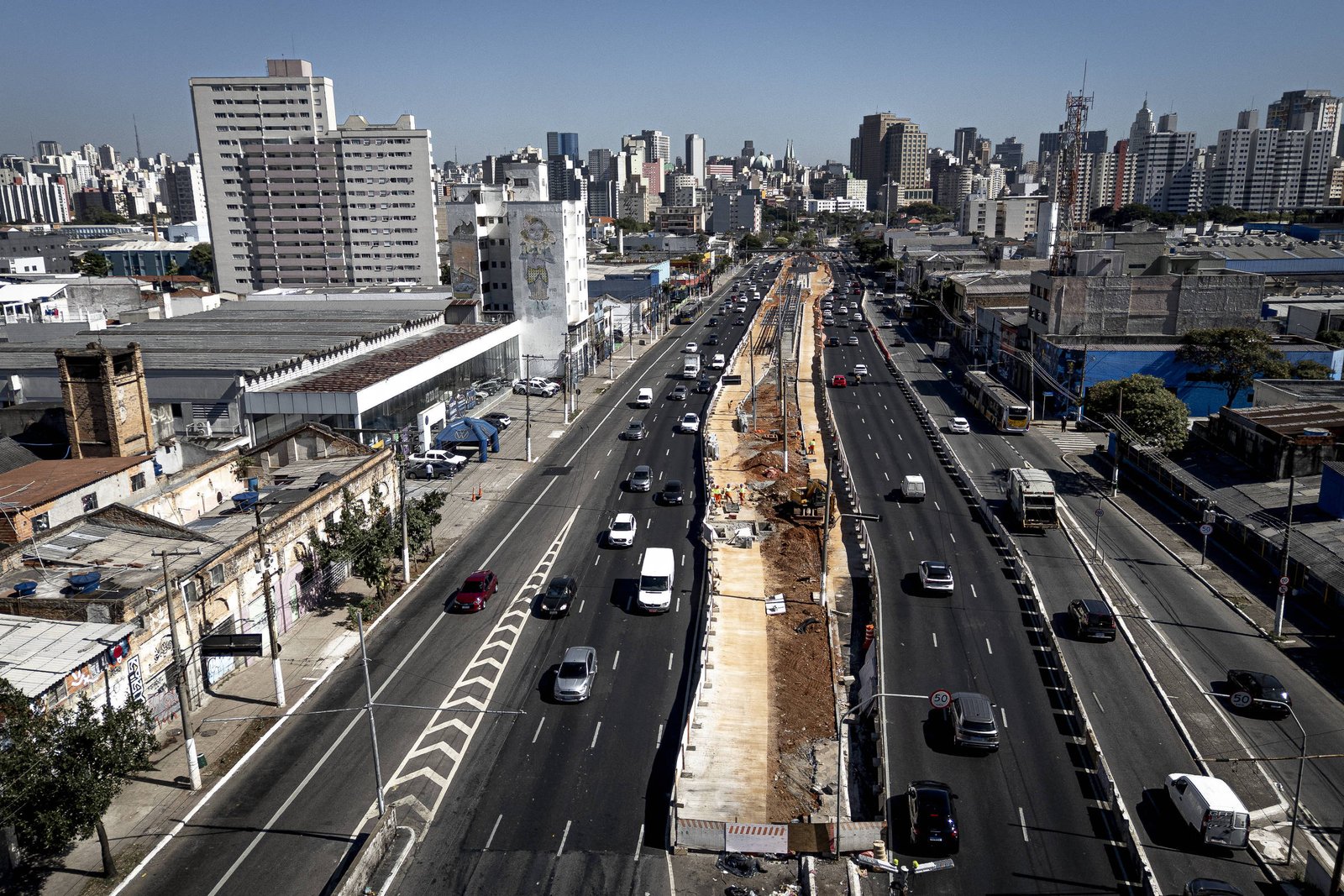 Corredor de ônibus em construção no canteiro central da Radial Leste com veículos circulando nas pistas laterais. Edifícios altos e céu claro ao fundo.