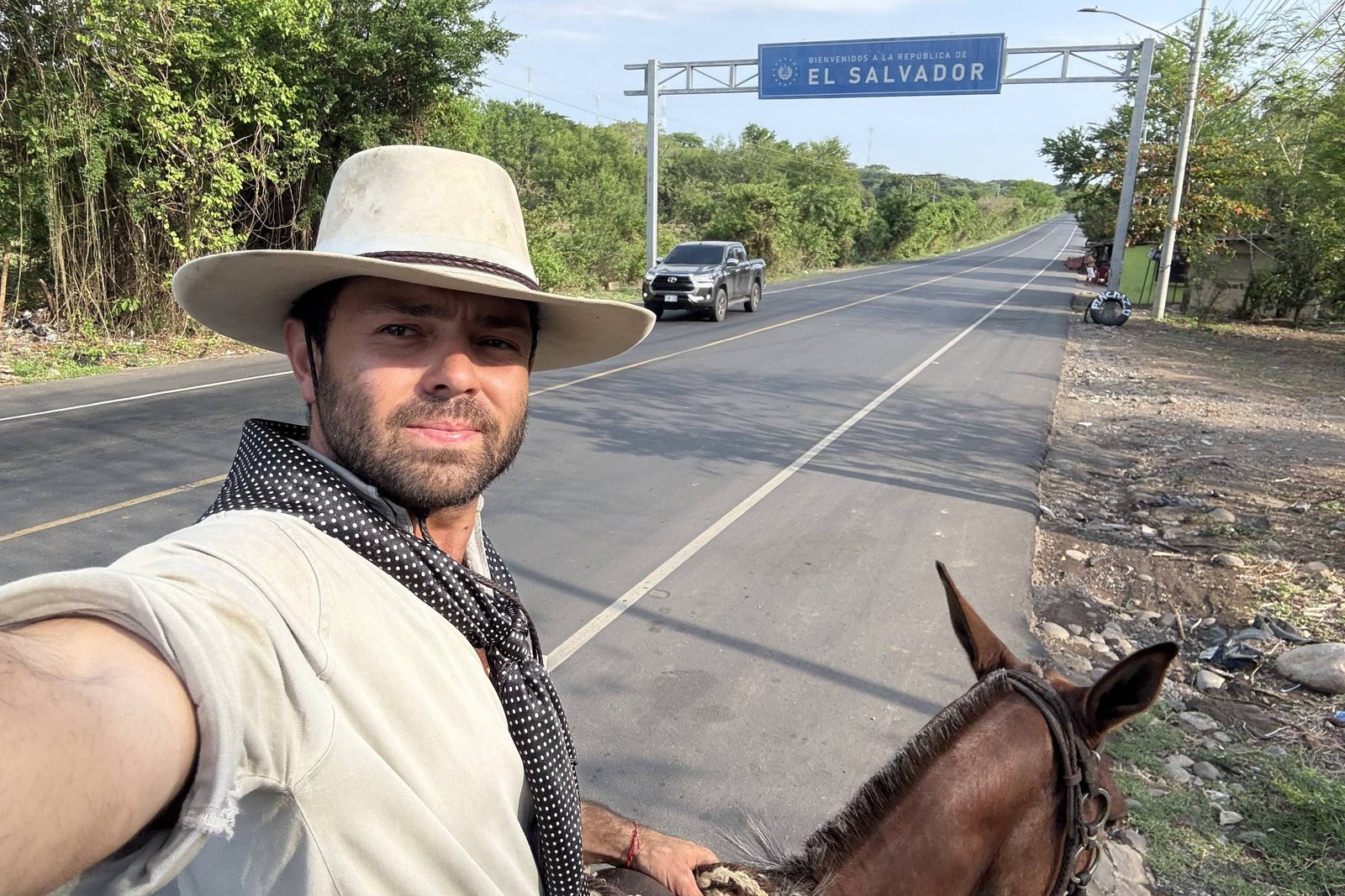 Homem com chapéu claro e lenço preto faz selfie montado em cavalo ao lado de estrada asfaltada. Placa azul indica entrada para El Salvador. Vegetação e céu claro ao fundo.