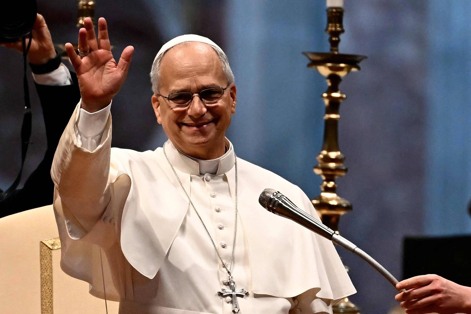 Homem vestido com vestes papais brancas, usando óculos e cruz pendurada no pescoço, acena com a mão direita em ambiente interno com candelabro ao fundo.