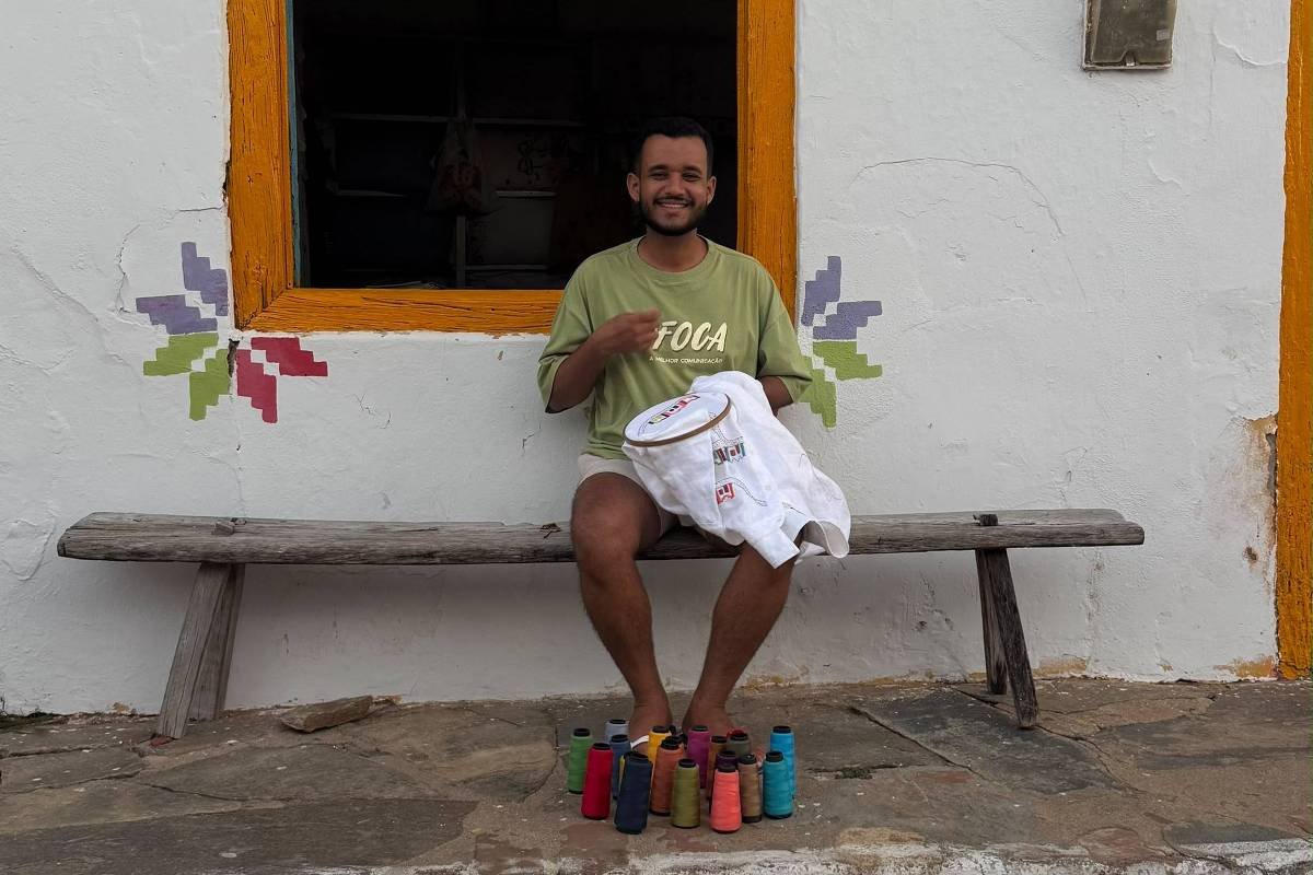 Homem sorridente sentado em banco de madeira em frente a casa branca com moldura amarela na janela. A janela tem grades pretas e é decorada com flores coloridas nas bordas. O homem segura uma camiseta branca e tem latas coloridas no chão à sua frente.