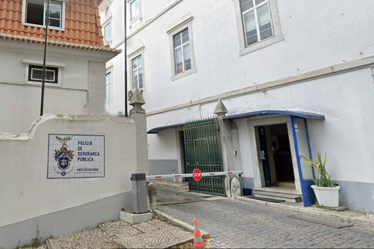 Entrada de prédio branco com portão verde aberto e placa na parede indicando Polícia de Segurança Pública. Calçada de pedra portuguesa e cone de sinalização laranja no chão.