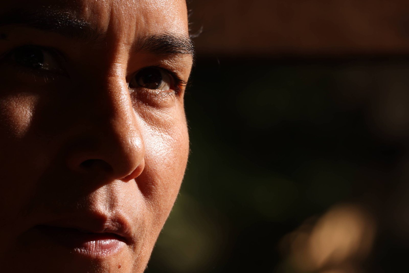 Close no rosto de uma mulher com pele morena, iluminada lateralmente, destacando olhos, nariz e boca. Fundo escuro desfocado.