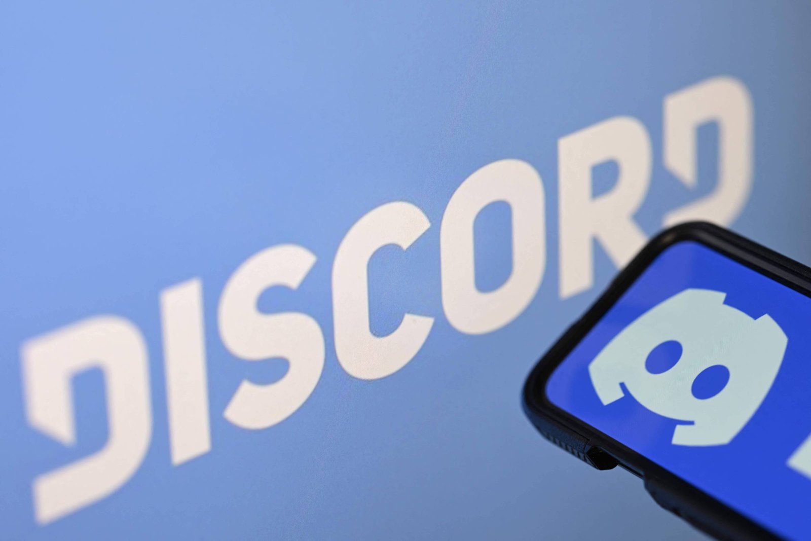 Tela de smartphone exibe o ícone branco do Discord sobre fundo azul. Ao fundo, a palavra