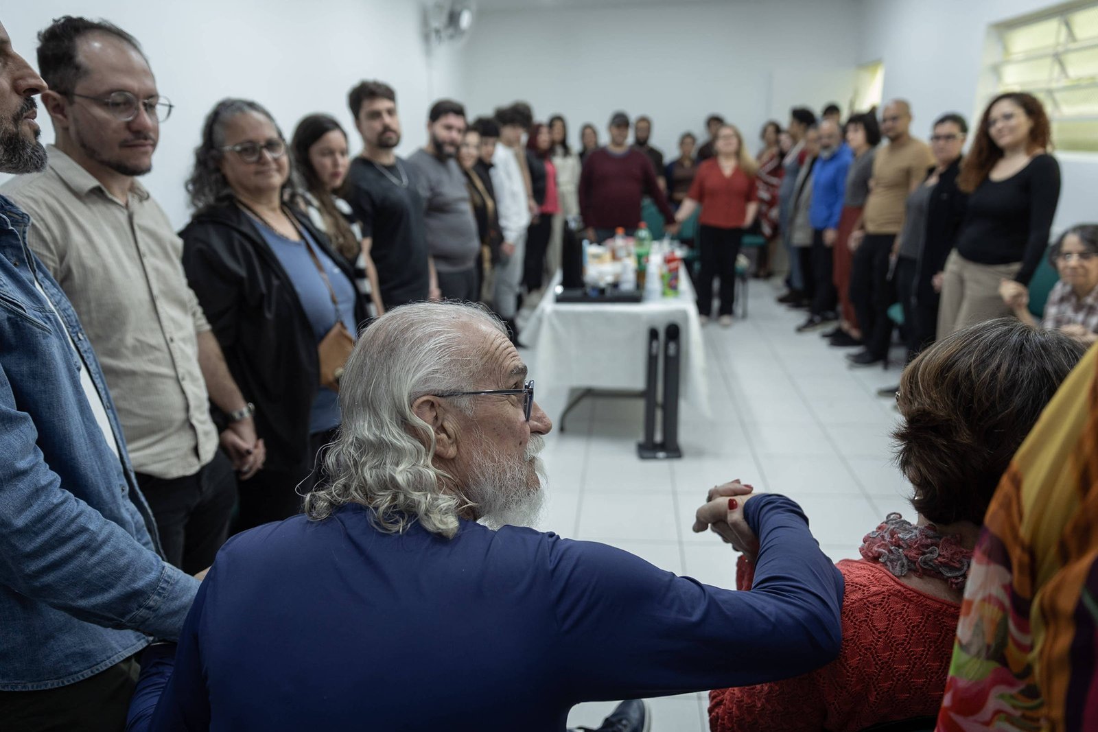 Cerca de 30 pessoas formam um círculo em sala branca com piso de azulejo. No centro, mesa com bebidas e copos. Homem de cabelos grisalhos e óculos cumprimenta mulher à sua frente.