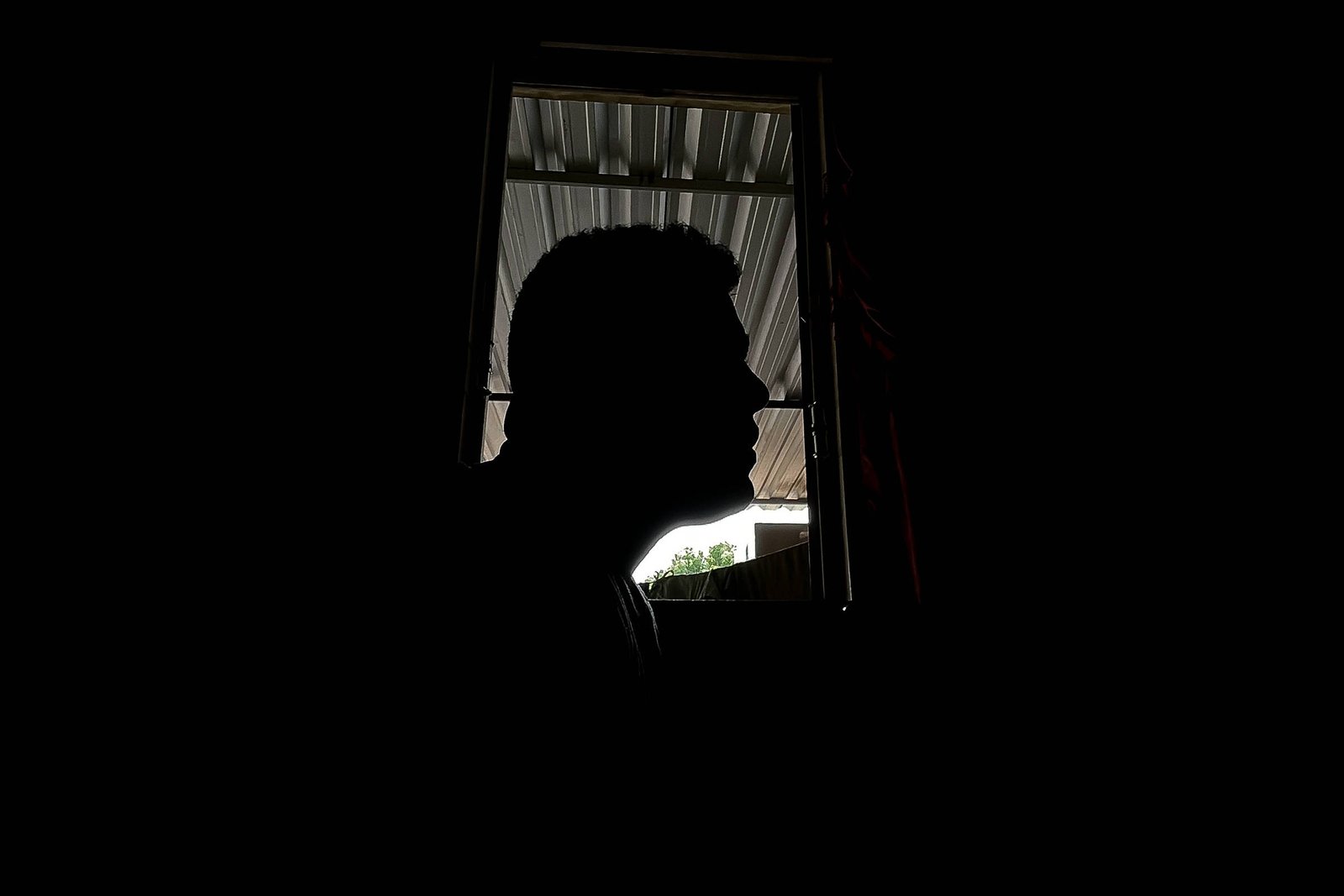 Silhueta de um homem de perfil vista contra a luz de uma janela aberta. Ao fundo, teto metálico e parte de área externa com vegetação visível.
