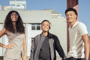 Três jovens adultos posam sorrindo em varanda de prédio durante dia ensolarado. À esquerda, mulher com cabelo cacheado e óculos, vestindo conjunto bege; ao centro, homem com jaqueta cinza e camiseta preta; à direita, homem com camiseta branca e calça preta, apoiado no parapeito. Edifícios residenciais ao fundo sob céu azul.