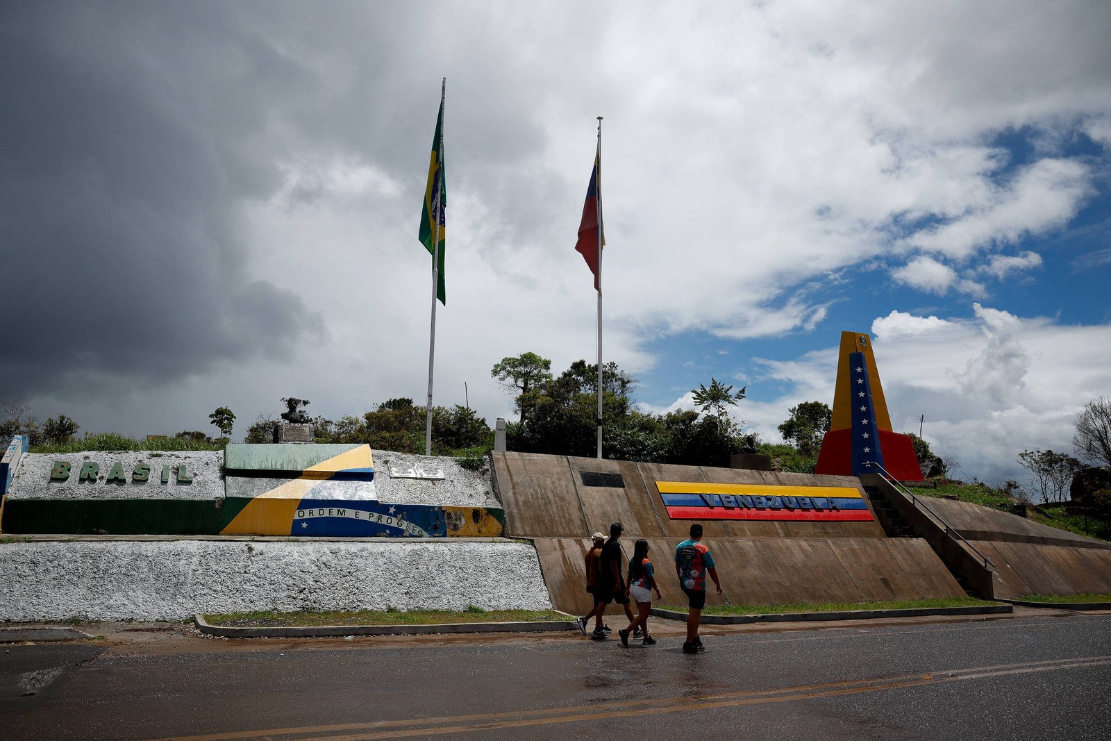 Marco fronteiriço com bandeiras do Brasil e da Venezuela hasteadas. Estrutura exibe as cores nacionais dos dois países e uma família caminha pela calçada sob céu parcialmente nublado.