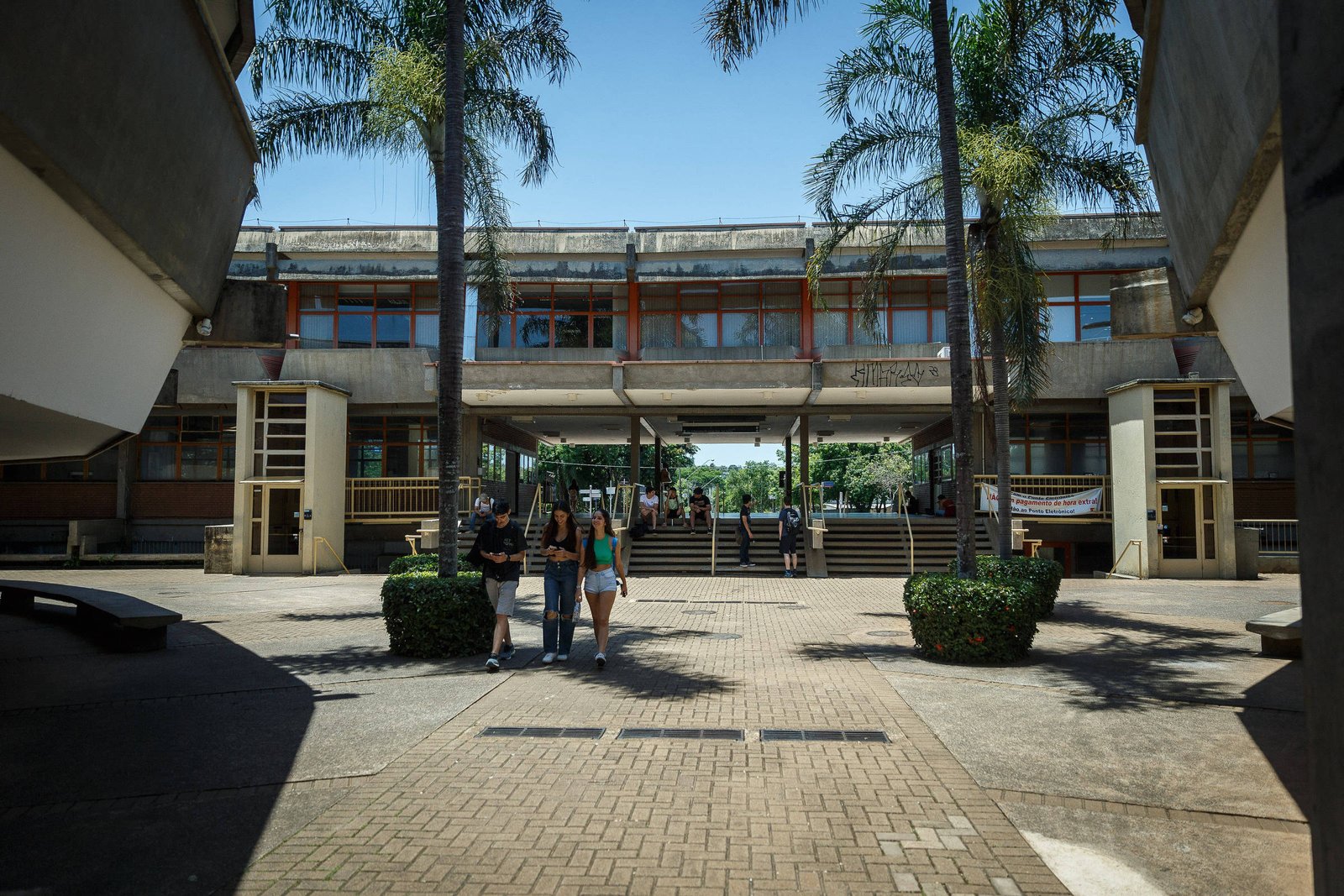 Vista da entrada principal de um campus universitário com três estudantes caminhando em direção à câmera. O prédio ao fundo tem estrutura de concreto e janelas largas, com palmeiras e arbustos decorativos ao redor do caminho de pedra. O céu está claro e azul, indicando dia ensolarado.