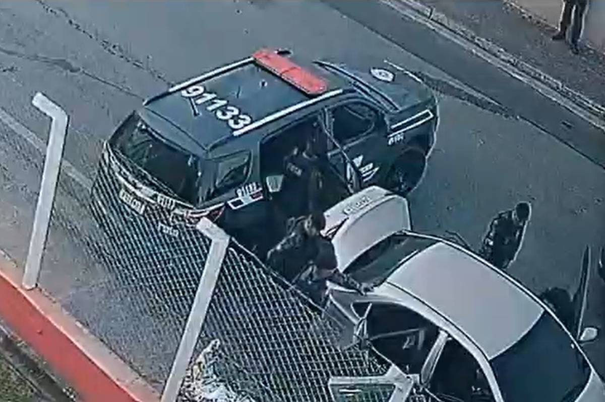 Uma viatura policial cinza está no centro da imagem, parcialmente sobre a calçada. Um carro prata está parado na via, com porta-malas aberto próximo à lateral do carro de polícia. É possível ver um homem entre a porta traseira da viatura da polícia e o carro prata, e outros dois policiais próximos ao porta-malas. Parte da cena é ocupada por uma carca branca de alambrado que encobre parcialmente a trasiera da viatura e os policiais