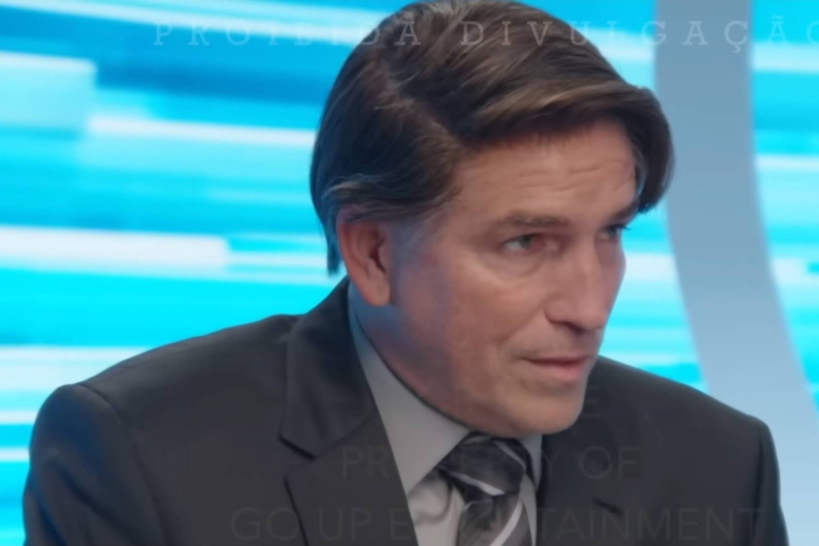 Homem de meia-idade com cabelo castanho escuro penteado para o lado, veste terno preto, camisa cinza e gravata cinza, sentado em estúdio com fundo azul claro e branco.