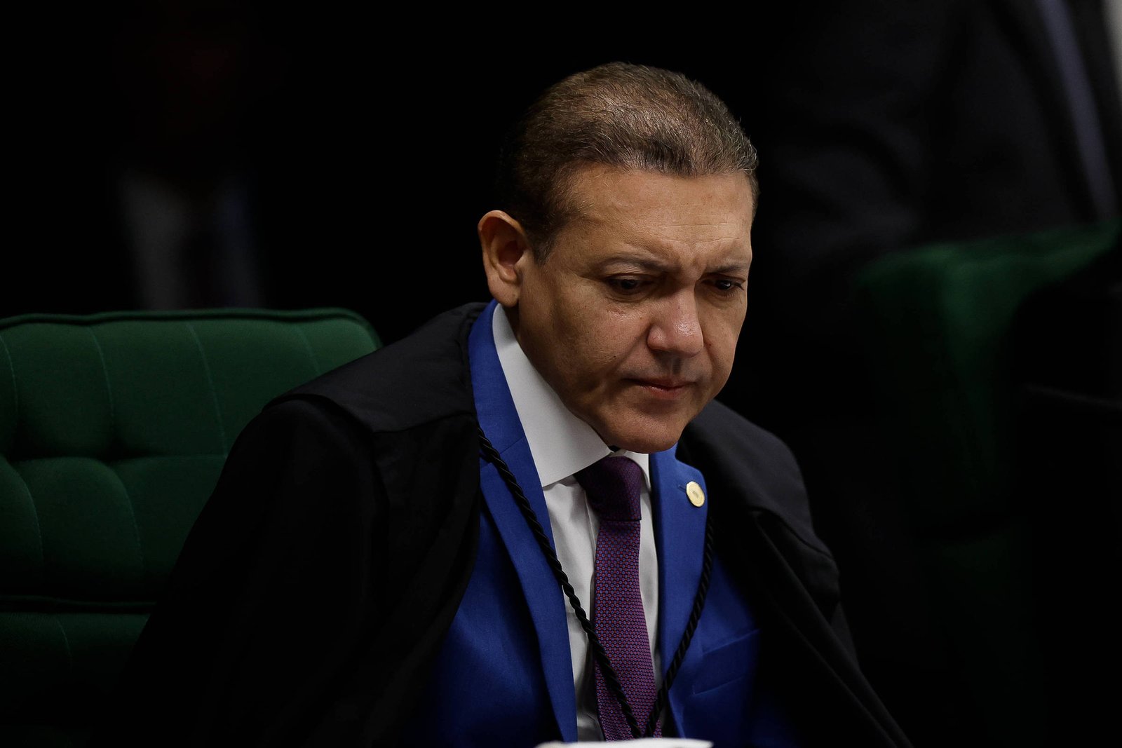 Homem de meia-idade com terno azul, gravata roxa e capa preta sentado em cadeira verde, com expressão séria, em ambiente escuro e formal.