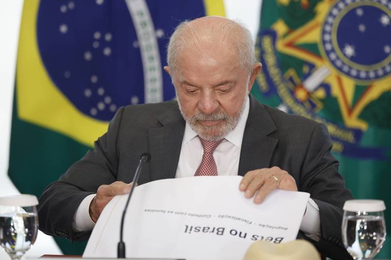 Homem de cabelos grisalhos e barba branca, vestido com terno escuro, camisa branca e gravata vermelha, sentado em mesa revisando documento intitulado 'Bens no Brasil'. Ao fundo, duas bandeiras do Brasil parcialmente visíveis. Na mesa, dois copos de água e microfone centralizado.