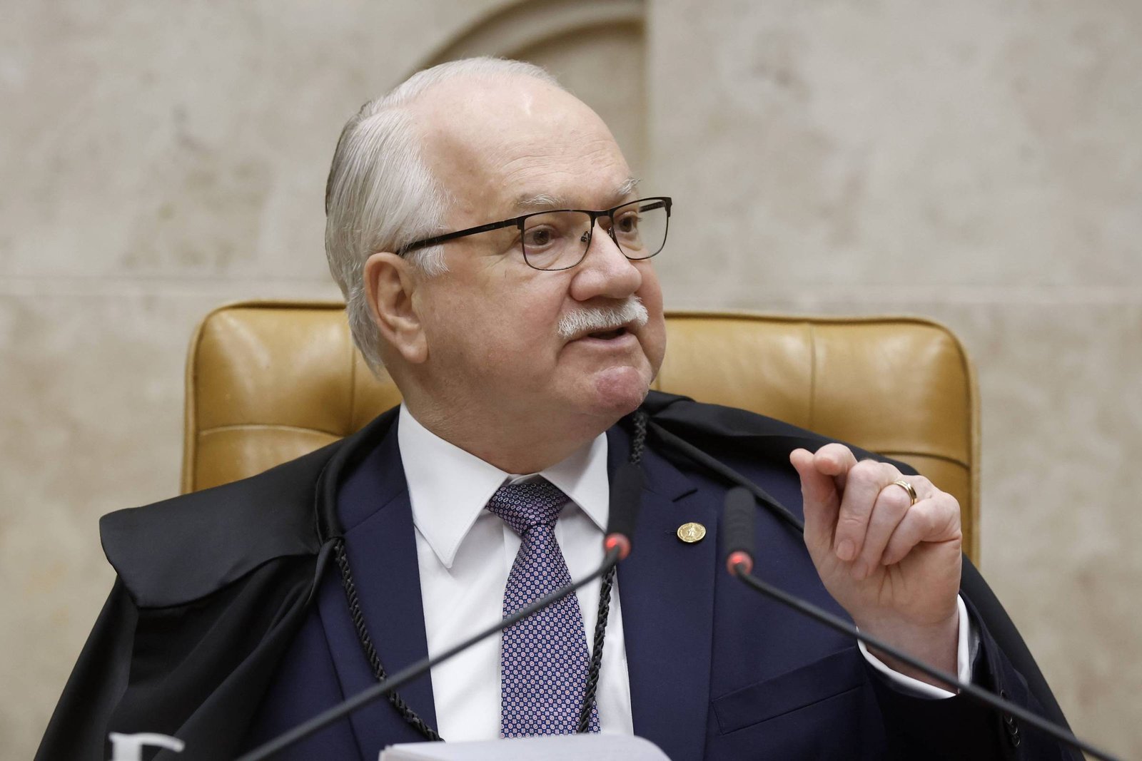 Homem idoso com cabelo branco e óculos, vestido com terno escuro, gravata e toga preta, sentado em cadeira de couro marrom, gesticulando com a mão direita enquanto fala em microfone.