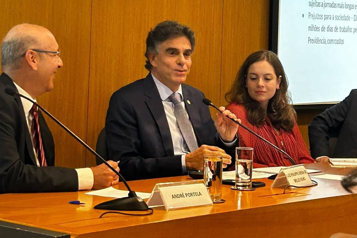 Três pessoas sentadas em mesa de conferência com microfones, copos d