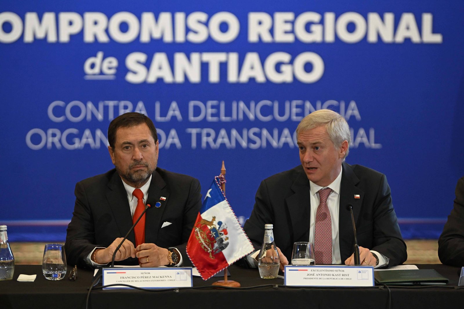 Dois homens sentados à mesa em conferência com microfones e bandeira do Chile ao centro. Fundo azul com texto