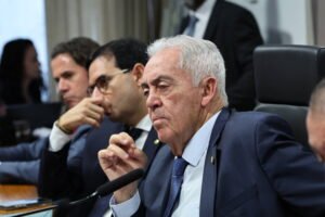 Homem idoso de cabelos brancos veste terno azul escuro e gravata azul, sentado em cadeira preta, participando de reunião formal. Ao fundo, outros três homens também vestidos formalmente estão atentos, um deles com óculos e mão no rosto.