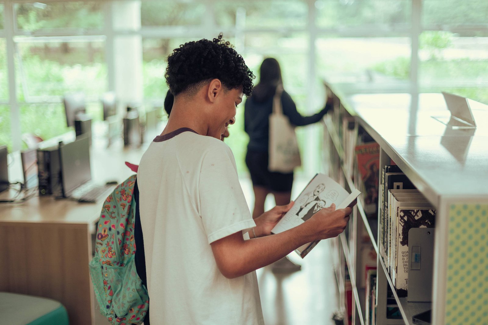Jovem com camiseta branca e mochila colorida lê uma revista em uma estante de biblioteca. Ao fundo, outra pessoa consulta livros em outra estante próxima a janelas amplas.