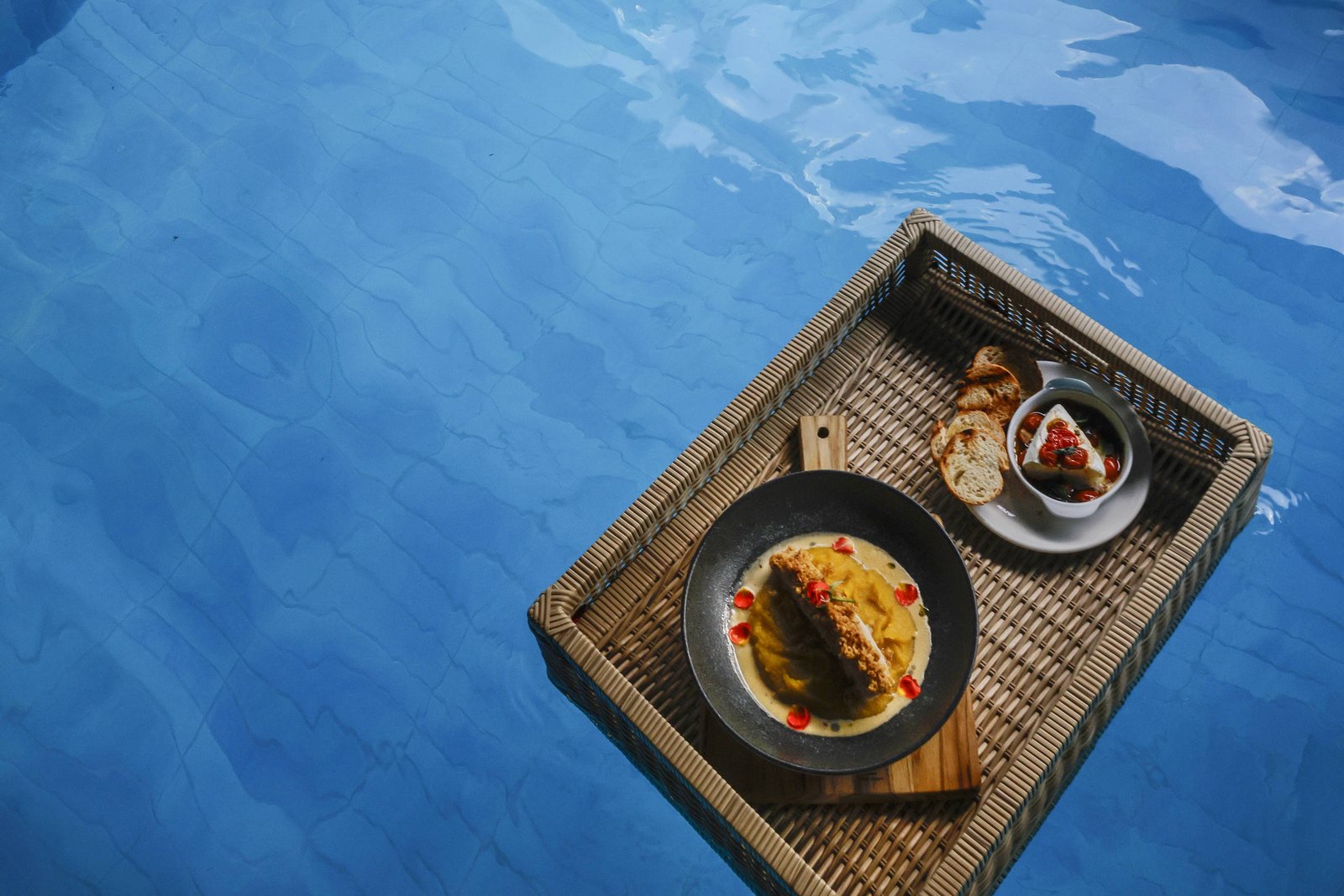 Bóia inflável branca em formato de cisne flutua em piscina de azulejos azuis. Bandeja de vime com prato de comida e bebida repousa na água. Parte do corpo de uma pessoa está visível na água ao lado.