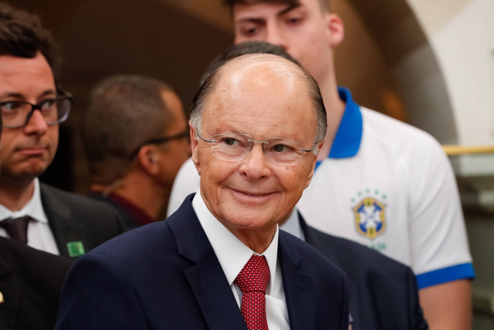 Homem de meia-idade com terno azul escuro, camisa branca e gravata vermelha sorri levemente. Ele usa óculos e está cercado por outras pessoas, incluindo um jovem com camisa branca e detalhes azuis com brasão do Brasil.