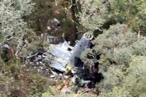 Destroços de uma aeronave parcialmente destruída estão entre árvores densas em área de mata fechada. Fragmentos metálicos e partes da fuselagem são visíveis no chão, sem presença de pessoas.