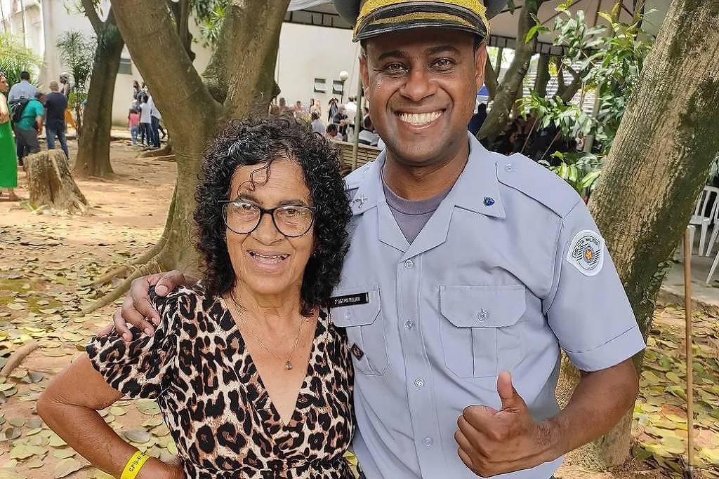 Homem vestido com uniforme cinza de policial militar sorri e faz sinal de positivo com a mão direita, enquanto abraça mulher de vestido estampado de oncinha e óculos. Eles estão em área externa com árvores e folhas no chão, com outras pessoas ao fundo.