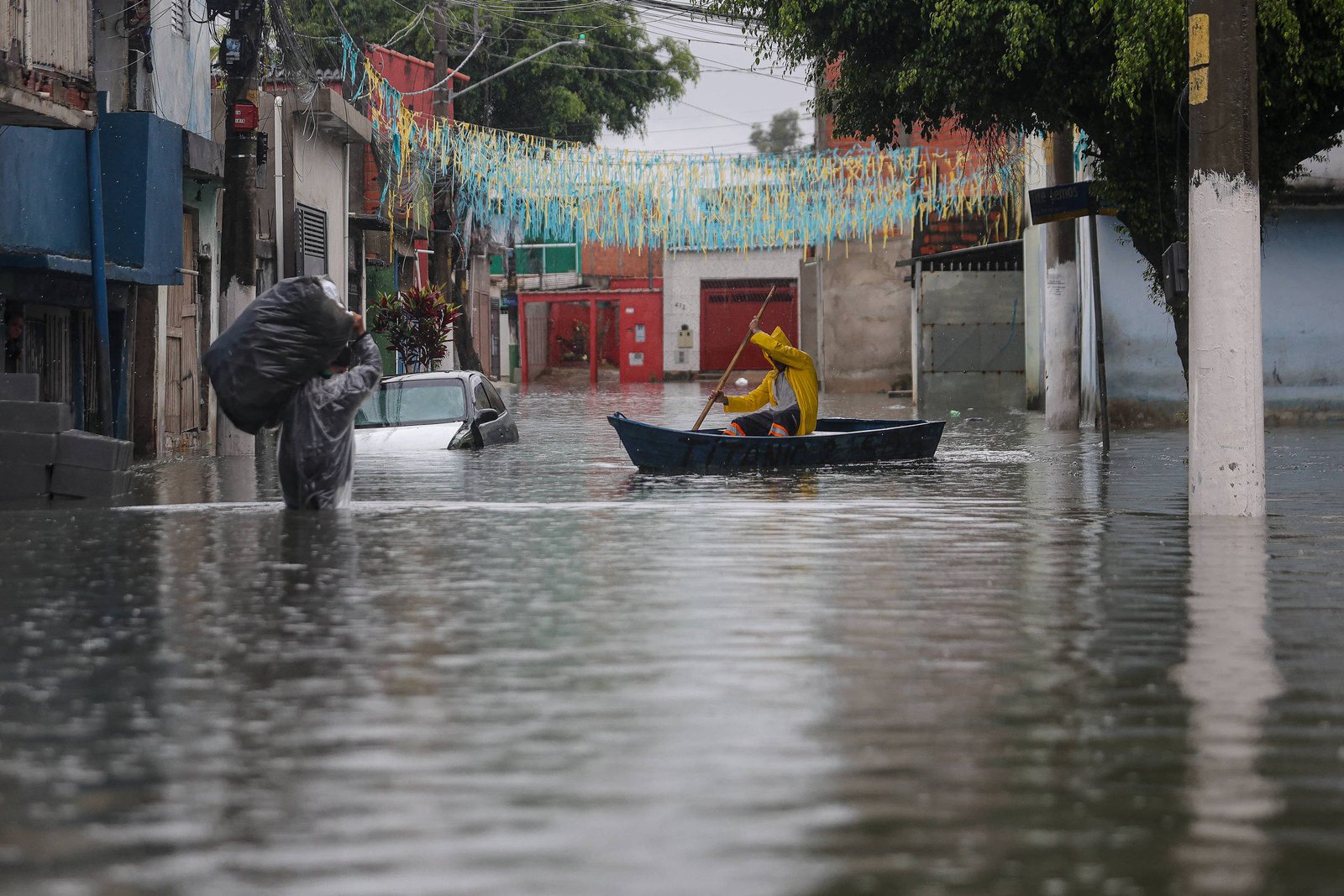 Rua residencial inundada com água na altura do peito. Pessoa à esquerda carrega saco grande na cabeça e outra pessoa à direita remando em barco pequeno. Casas e carros parcialmente submersos, decoração colorida pendurada acima da rua.