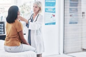 Médica de jaleco branco examina pescoço de paciente sentada em maca dentro de consultório. Ambiente claro com cartazes informativos na parede.