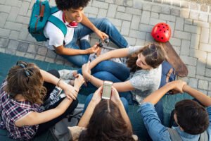 Um grupo de cinco jovens sentados em um espaço ao ar livre, em um piso de pedras. Eles estão em uma posição descontraída, com alguns segurando celulares e outros interagindo entre si. Um jovem com cabelo cacheado e mochila está no centro, enquanto uma jovem com cabelo longo e liso está olhando para um celular. Um skate e um capacete vermelho estão próximos ao grupo.