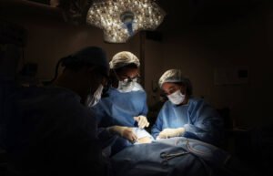 A imagem mostra uma equipe cirúrgica composta por três profissionais em um ambiente de cirurgia. Eles estão usando roupas cirúrgicas, incluindo toucas e máscaras. A iluminação é focada na área de operação, destacando o procedimento que está sendo realizado. O fundo é escuro, o que contrasta com a luz intensa do equipamento cirúrgico acima.