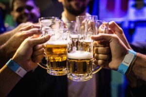 Quatro pessoas erguem canecas de cerveja dourada com espuma em brinde. Duas delas usam pulseiras azuis. Fundo desfocado indica ambiente interno com iluminação azul e roxa.