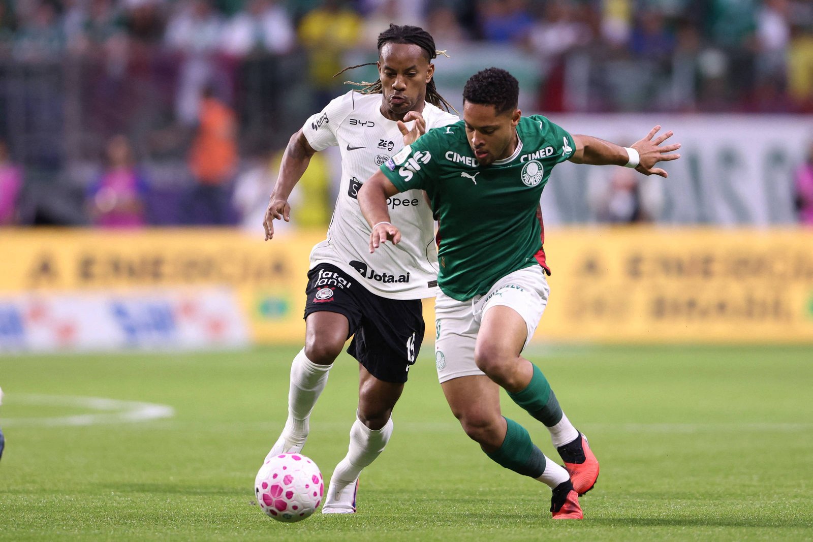 Dois jogadores disputam a posse da bola em campo de futebol. Um veste uniforme verde e branco, o outro branco e preto. O jogador de verde tenta avançar com a bola enquanto o adversário tenta impedir.