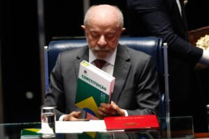 Homem idoso de terno cinza e gravata vermelha está sentado em cadeira azul, segurando um exemplar da Constituição brasileira com capa verde, amarela e branca. Há um copo de água e um documento vermelho sobre a mesa de vidro à sua frente. Fundo escuro e outra pessoa parcialmente visível ao fundo.