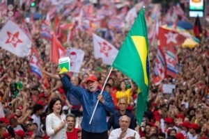 Homem no centro segura bandeira do Brasil e um livro, cercado por multidão com bandeiras vermelhas e símbolos políticos. Pessoas vestem vermelho e aplaudem em ambiente urbano.