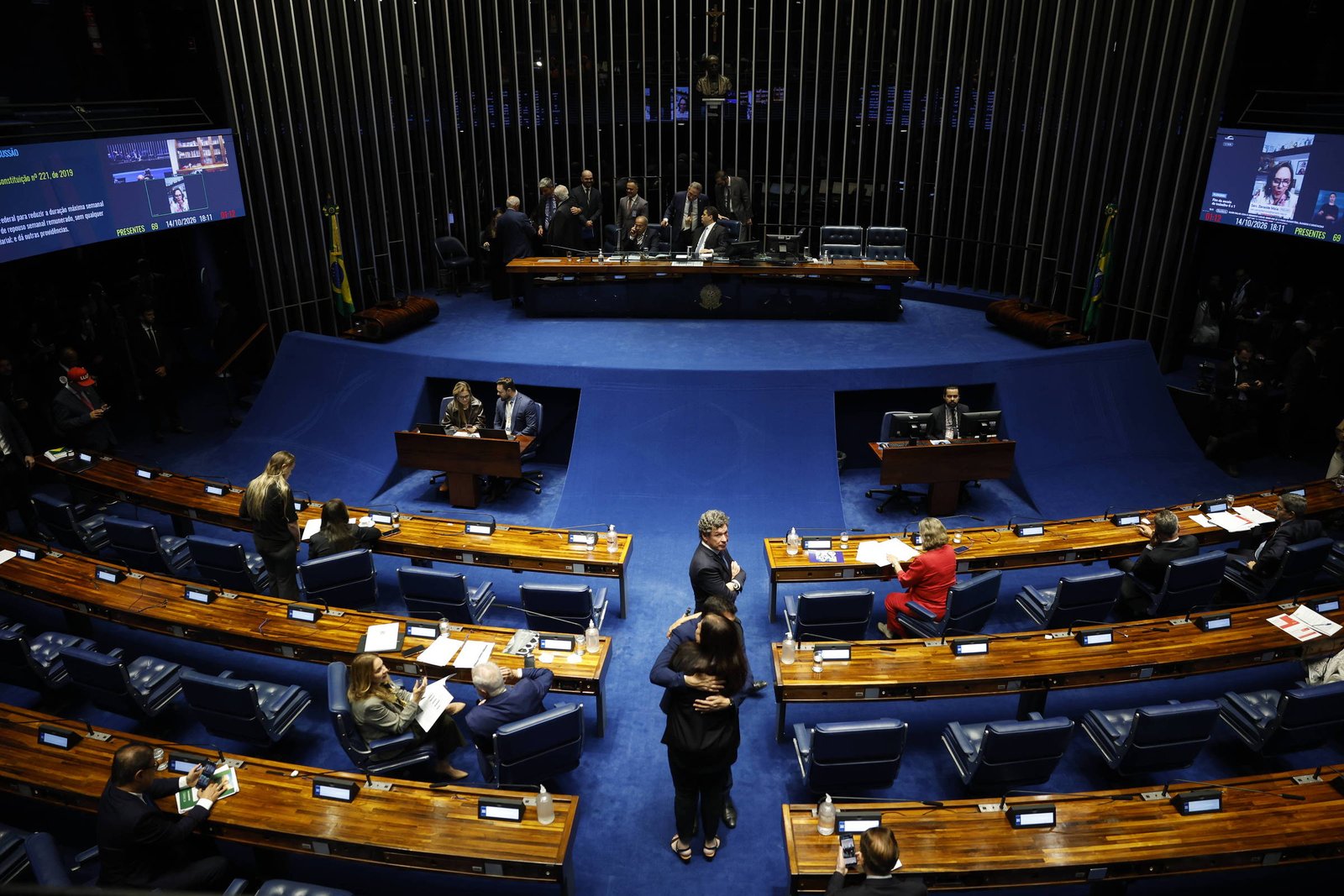 Plenário do Senado Federal com poucos senadores sentados em cadeiras azuis e mesas de madeira. No centro, duas pessoas conversam em pé. Ao fundo, mesa da presidência com cinco pessoas sentadas. Telas eletrônicas exibem informações nas laterais da sala.