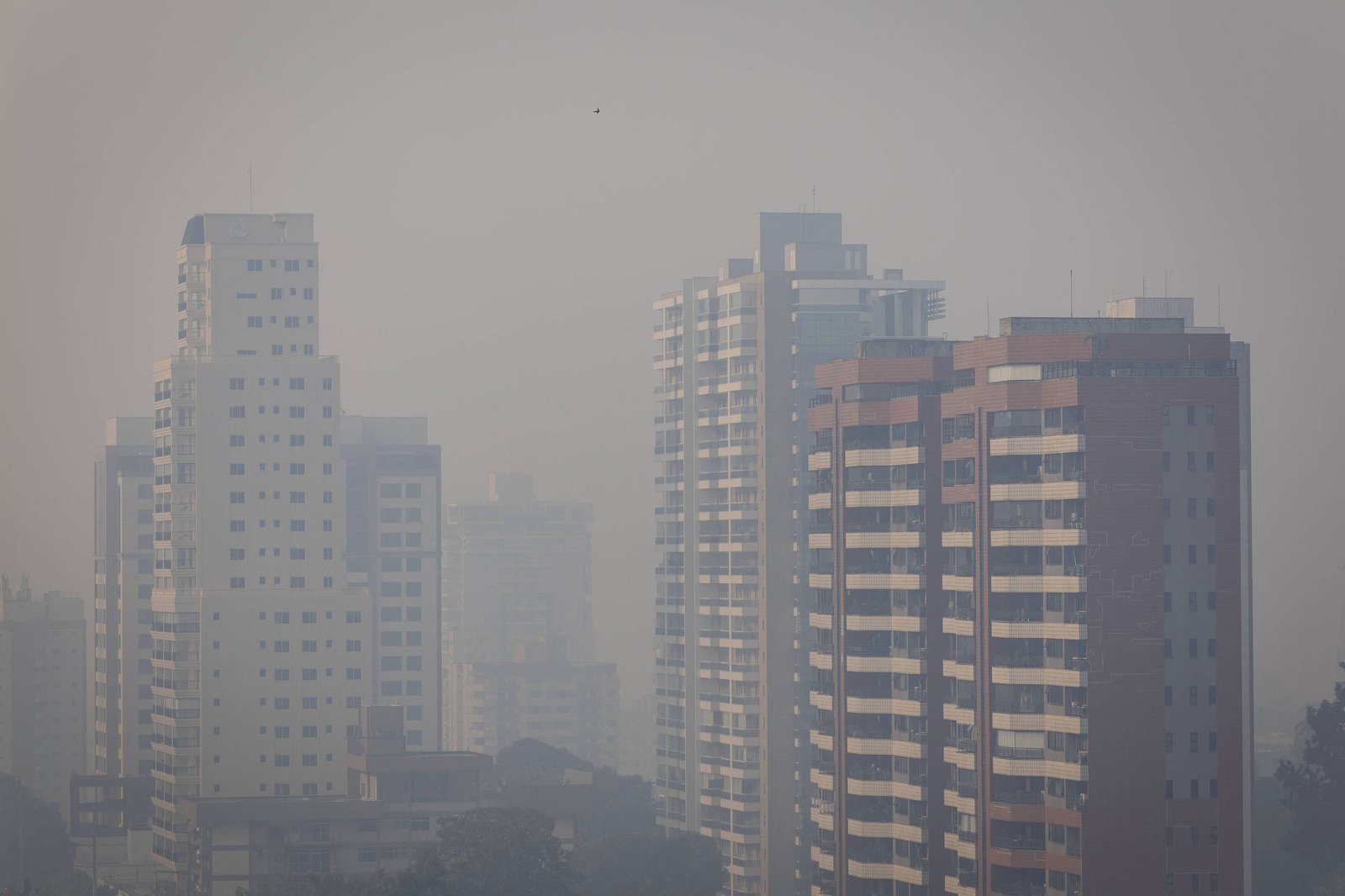 Vários prédios residenciais altos em área urbana sob forte neblina ou poluição, reduzindo a visibilidade. O céu está cinza e encoberto, dificultando a distinção dos edifícios ao fundo.