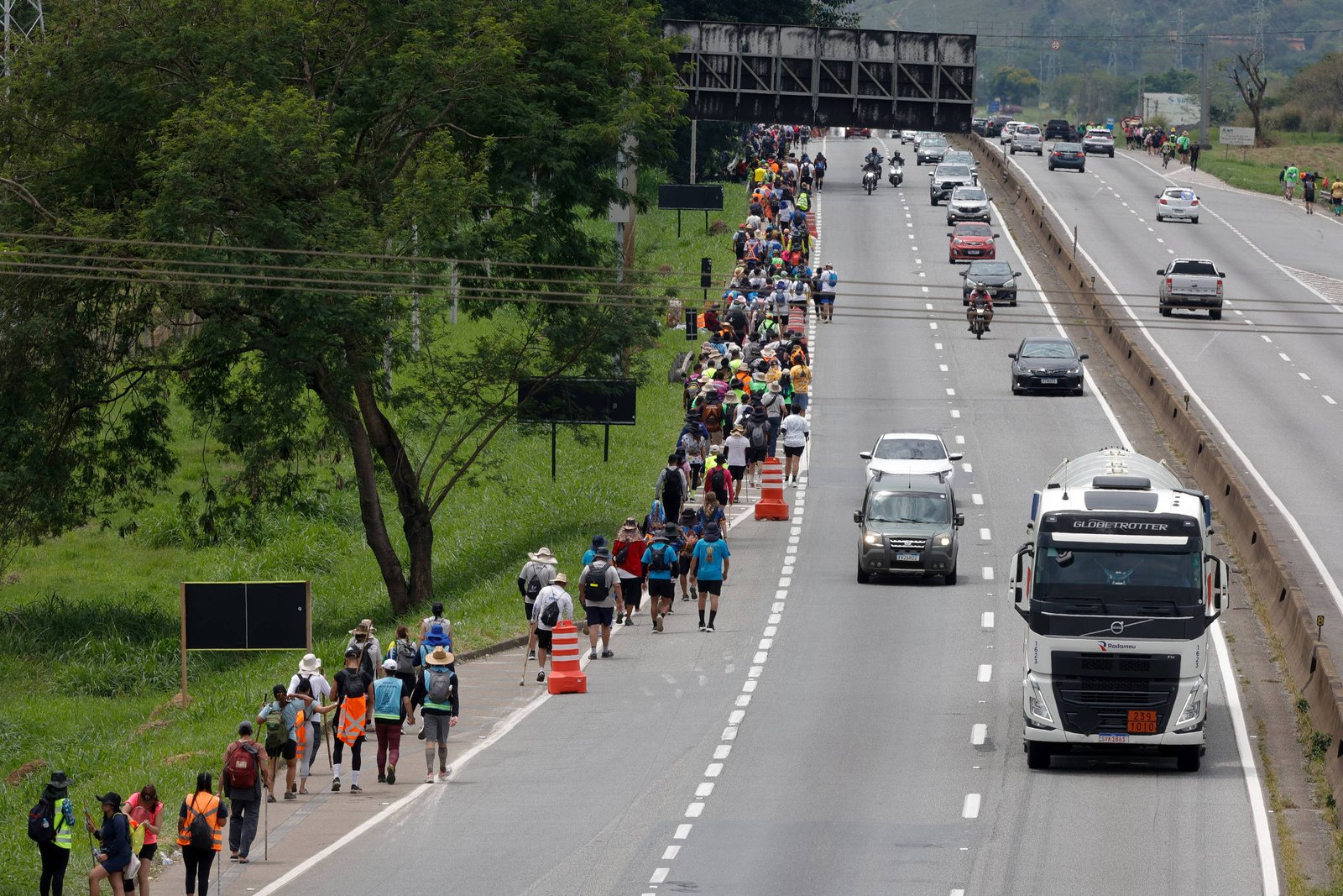 Centenas de pessoas caminham em fila ao longo do acostamento de uma rodovia, acompanhadas por cones laranja que delimitam a área. Veículos, incluindo caminhões e carros, trafegam nas faixas ao lado. Vegetação verde e árvores estão presentes à esquerda da via.