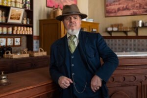 Homem de barba branca usa terno azul escuro, colete, gravata verde e chapéu marrom, apoiado no balcão de madeira de uma loja antiga. Ao fundo, prateleiras com potes, cestas e quadros nas paredes.