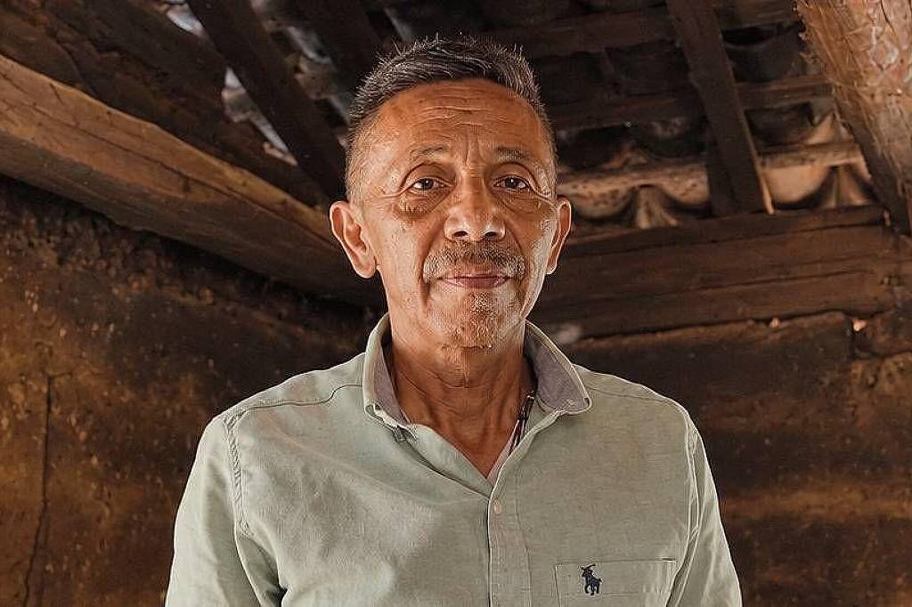 Homem de meia-idade com camisa clara de botão está em ambiente interno com paredes e teto de madeira rústica visíveis ao fundo.