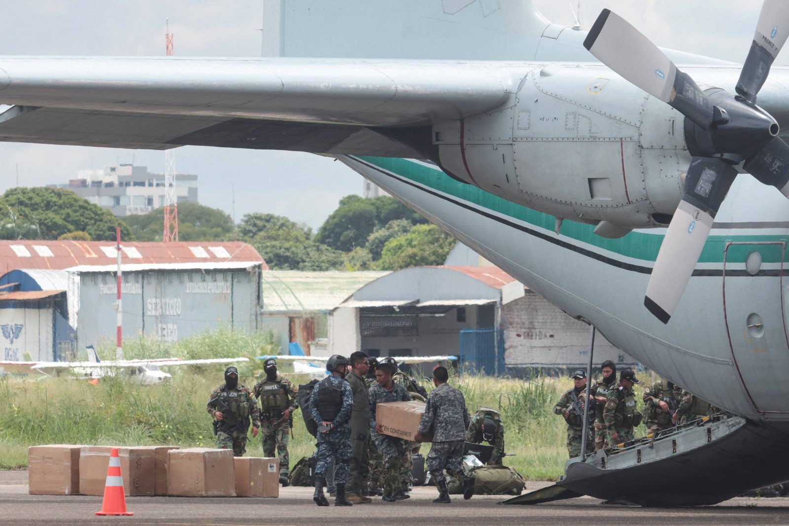 Grupo de militares em uniforme camuflado carregam caixas de papelão para dentro da área de carga de um avião de transporte estacionado em pista de aeroporto. Ao fundo, há hangares e vegetação.