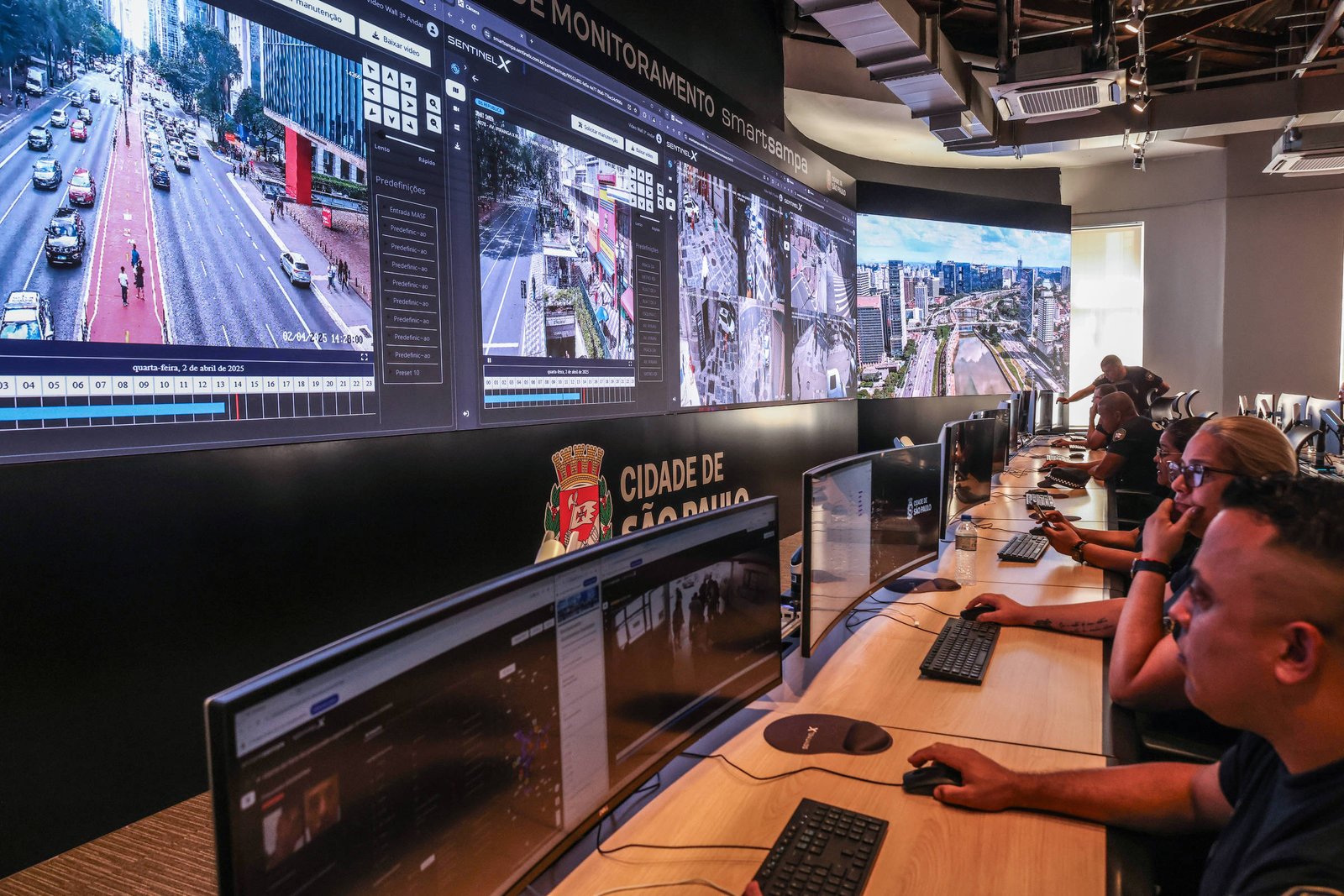 Sala com operadores sentados em frente a múltiplos monitores exibindo imagens de câmeras de segurança e tráfego da cidade de São Paulo. Na parede, grandes telas mostram vistas aéreas e de ruas movimentadas, com o brasão da cidade visível abaixo.