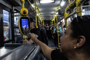 Passageira estende cartão para validar passagem em leitor eletrônico amarelo dentro de ônibus urbano lotado. Outros passageiros estão sentados e em pé ao fundo, segurando barras de apoio amarelas.